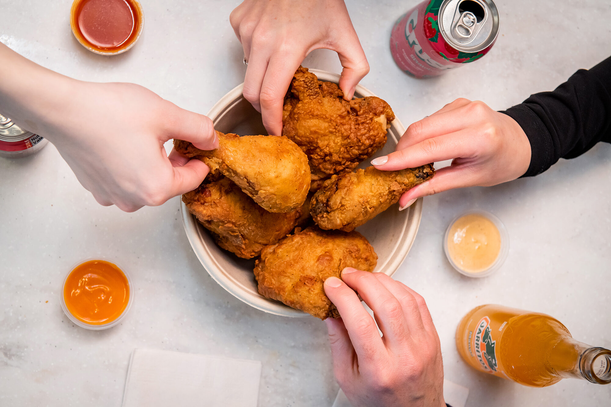 four hands grab pieces of friend chicken from a white paper bowl